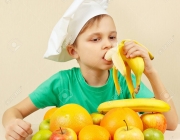 Little boy in chefs hat eat fresh banana at the table with fruits