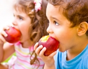 Photo of two happy children eating apples, brother and sister having picnic outdoors, cheerful kids biting red ripe peach, adorable infant holding fresh fruits in hands, healthy nutrition concept